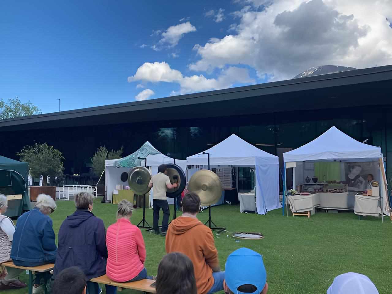 Open-Air-Performance mit Gongs vor weißen Marktzelten, Publikum sitzt auf der Wiese.